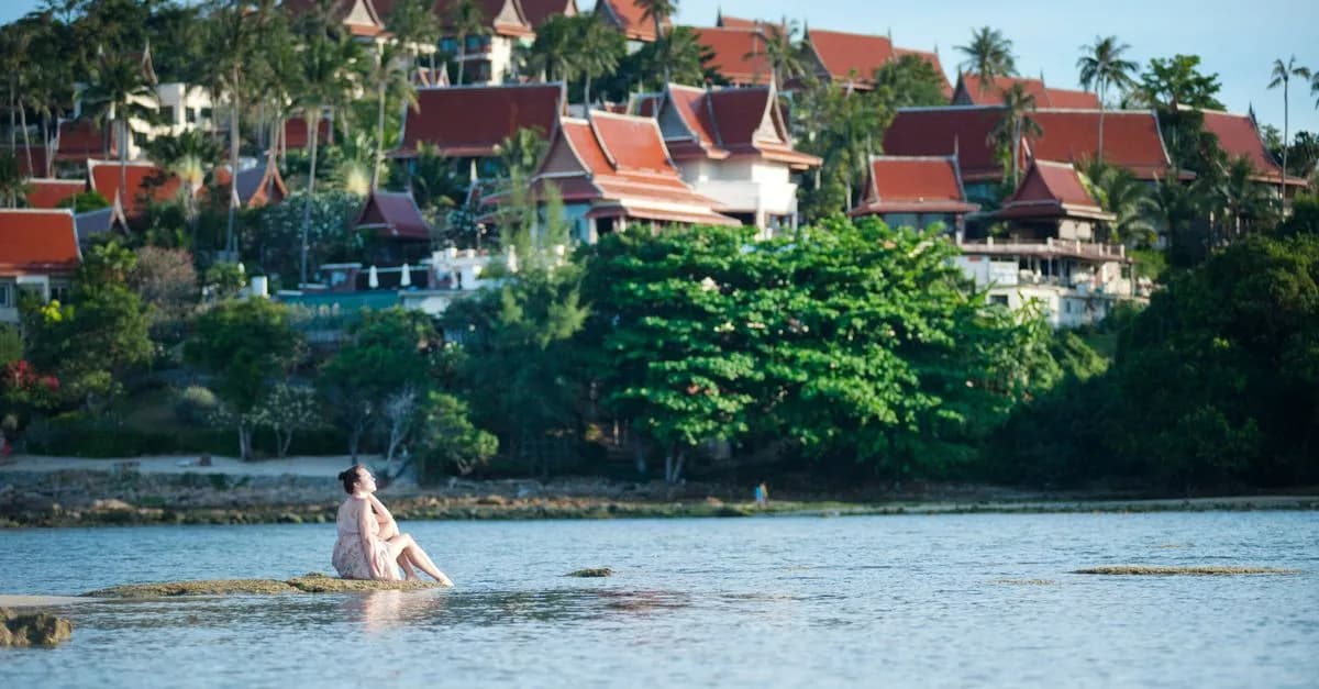 Woman relaxing by water with Thai resort in background.