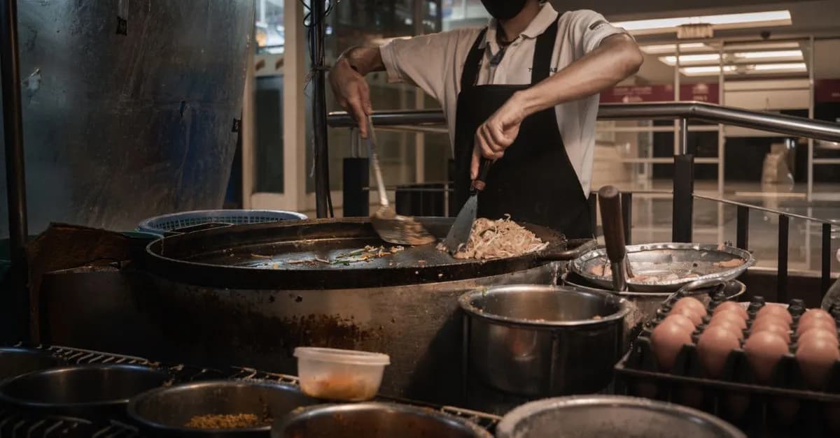 A masked chef skillfully prepares traditional street food in a bustling Bangkok night market.