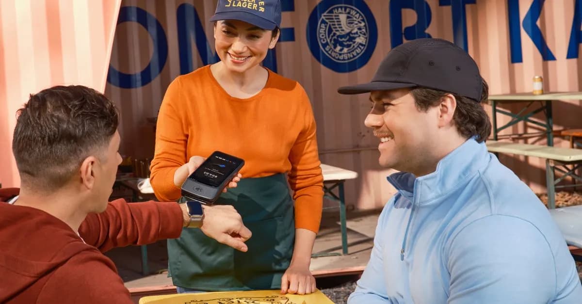 Friends using smartwatch for contactless payment at an outdoor cafe, assisted by a smiling waitress.