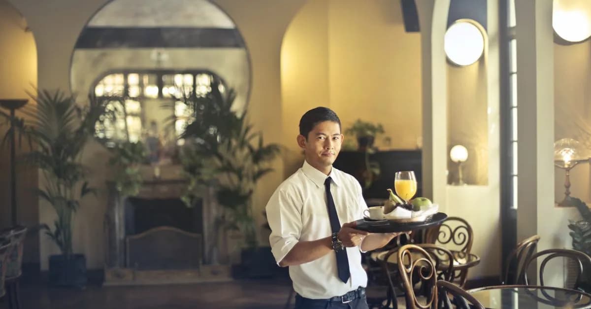 Confident young ethnic waiter in elegant clothes holding tray with food and drinks and looking at camera while serving tables in stylish restaurant