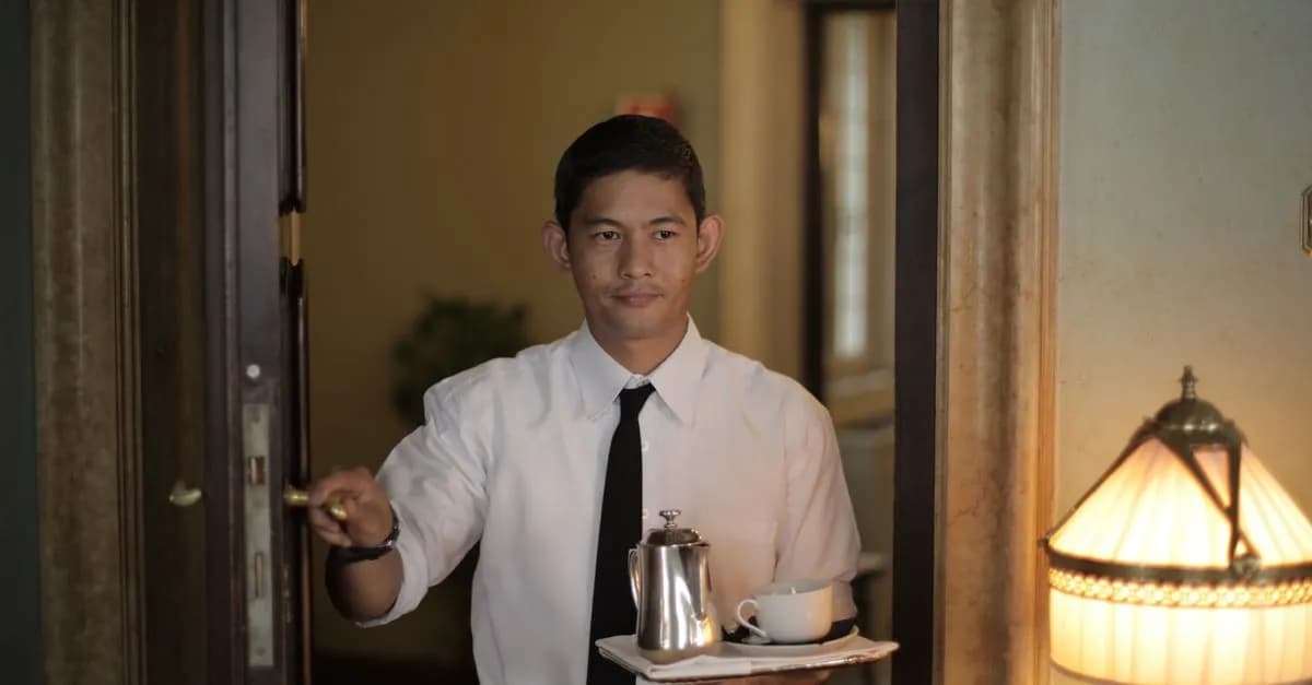 Positive ethnic male employee carrying tray with coffee while serving guest in cozy hotel room