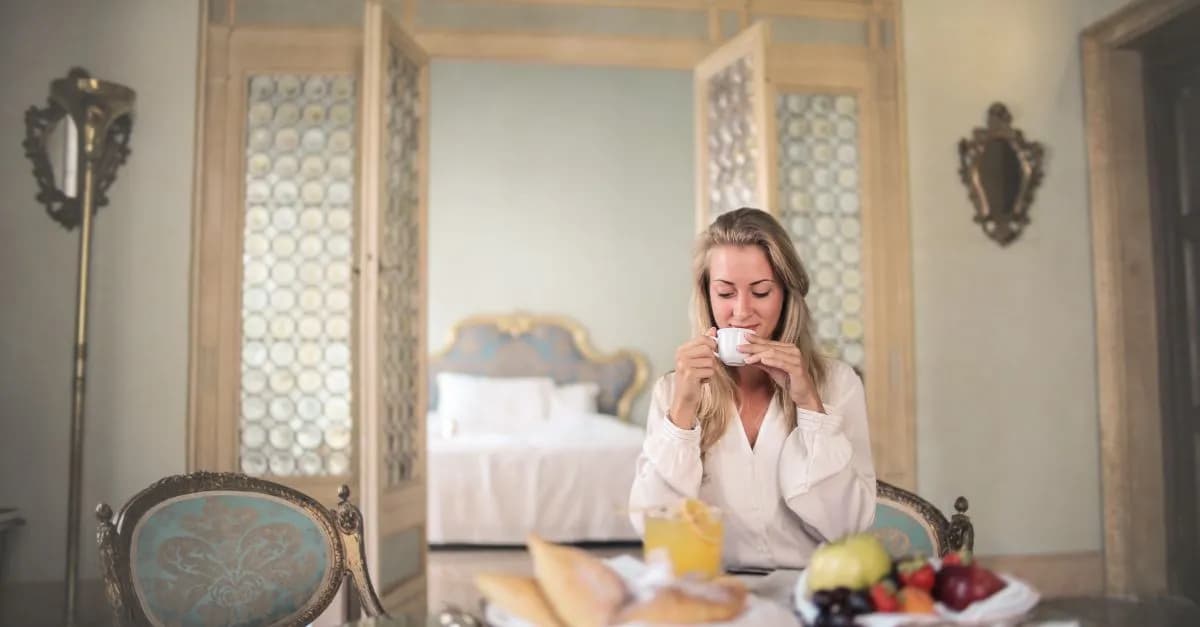 Glad female in elegant wear sitting at table with metal tray of healthy breakfast in vintage hotel room and drinking coffee in morning