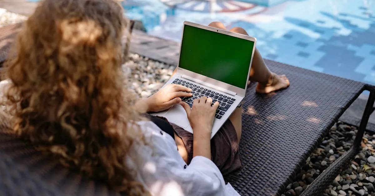 From above back view of anonymous female freelancer typing on portable computer while lying on sunbed near swimming pool with transparent water in sunlight