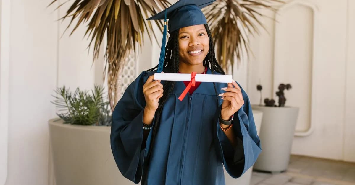 Young woman celebrating graduation with diploma in hand, wearing cap and gown, smiling proudly.