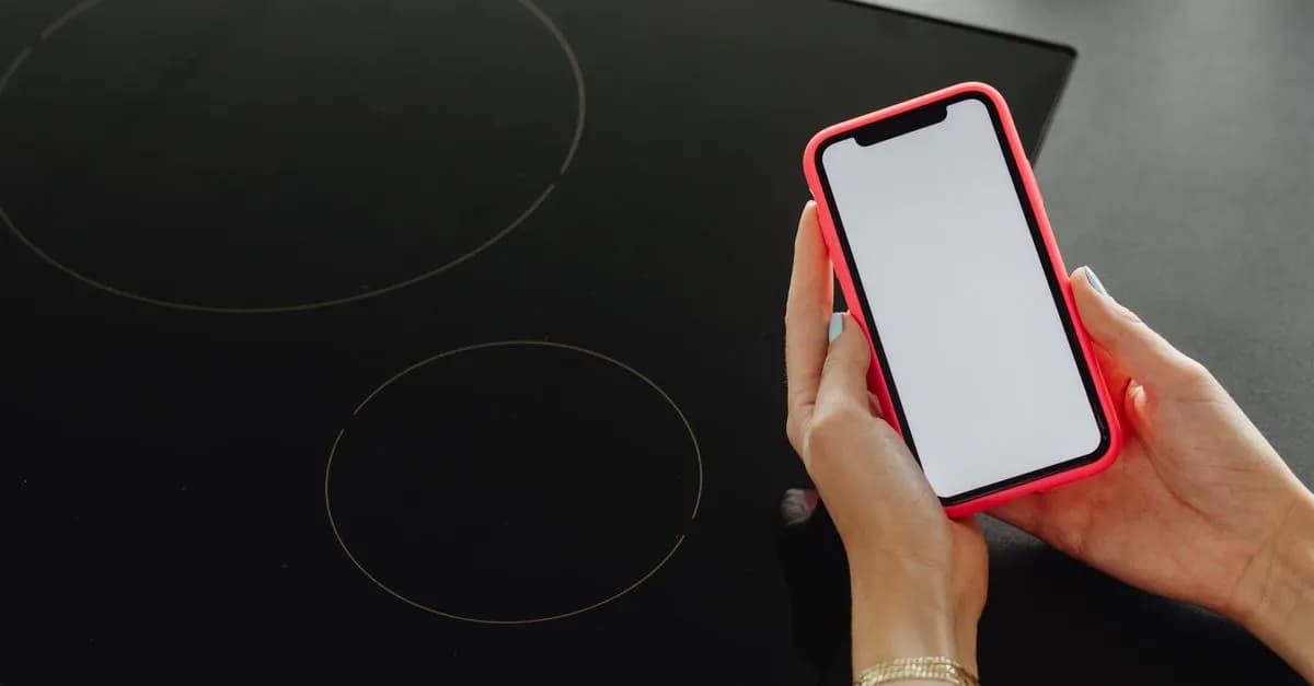 Close-up of hands holding a smartphone with a blank screen, beside a black cooktop.