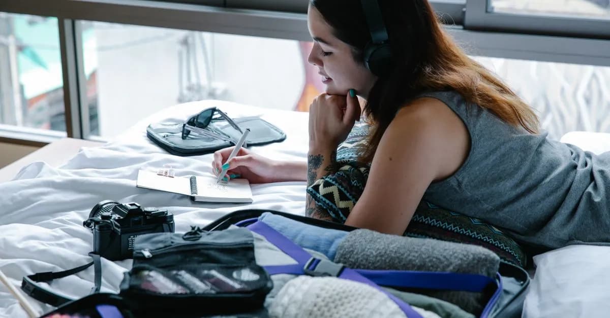 Woman lying on bed planning travel with suitcase, camera, and headphones.